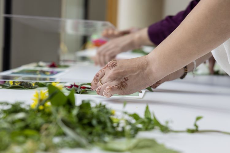 Close-up of hands pressing flowers and leaves onto paper during a creative activity. One person has floral henna designs on their hand and is wearing a gold ring.
