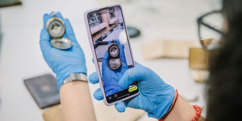 A pair of gloved hands. One hand holds an artifact and the other is takes a photo with a smartphone.