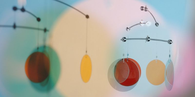 A mobile hangs in the foreground, consisting of a metal frame with coloured lenses suspended from it. Behind it, the shadow of the mobile and coloured circles and ellipses are projected onto a wall.