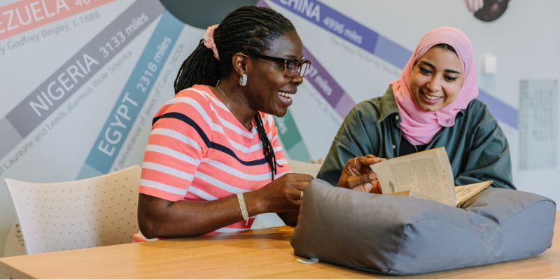 Two women are sat next to each other at a table. They are looking at, and turning the pages, of an old book that rests on a cushion. They are both smiling and look interested in the book.