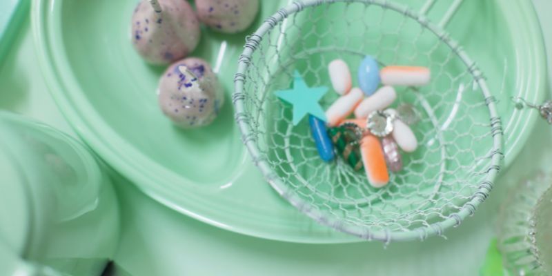 A pastel green hospital food tray on a pastel green table. In the left half of the tray are brown and purple spotted balls on sticks. In the right side of the tray is a wire basket filled with colourful pills.