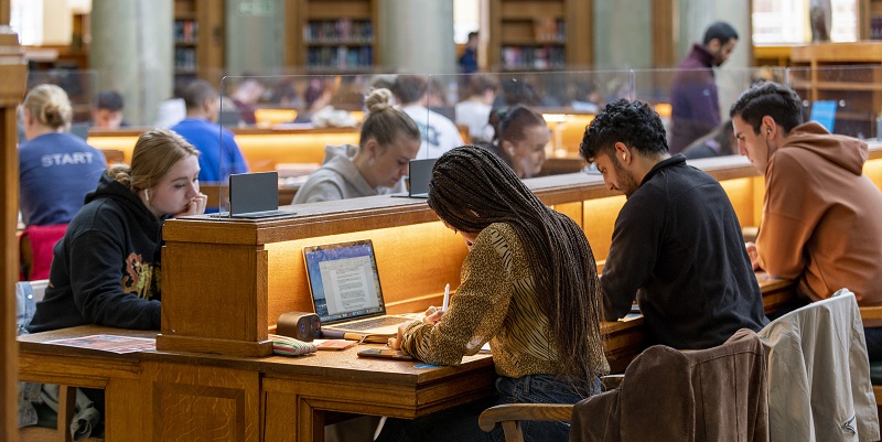 Students sit working with laptops at a long oak desk with task lighting in a busy Brotherton reading room.