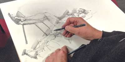 Photograph of hands holding a pencil and sketching a life drawing
