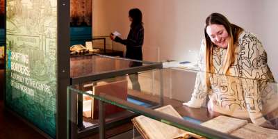 A gallery visitor looks at a museum case containing a historic travel guide book, next to a glowing plinth which says 'Shifting Borders'