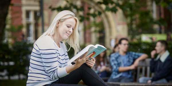 A student reads a book sat outdoors on campus.