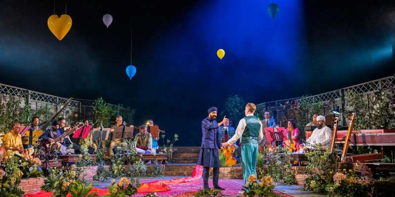 Photograph of two performers on a stage filled with flowers and musicians around the edge, against a dark blue backdrop.
